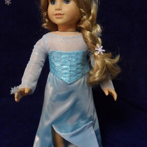 May include: A doll with blonde hair styled in a braid, wearing a light blue dress with a bodice and a slit in the skirt. The doll has blue eyes and is wearing silver shoes. Snowflake hair accessories complete the look.