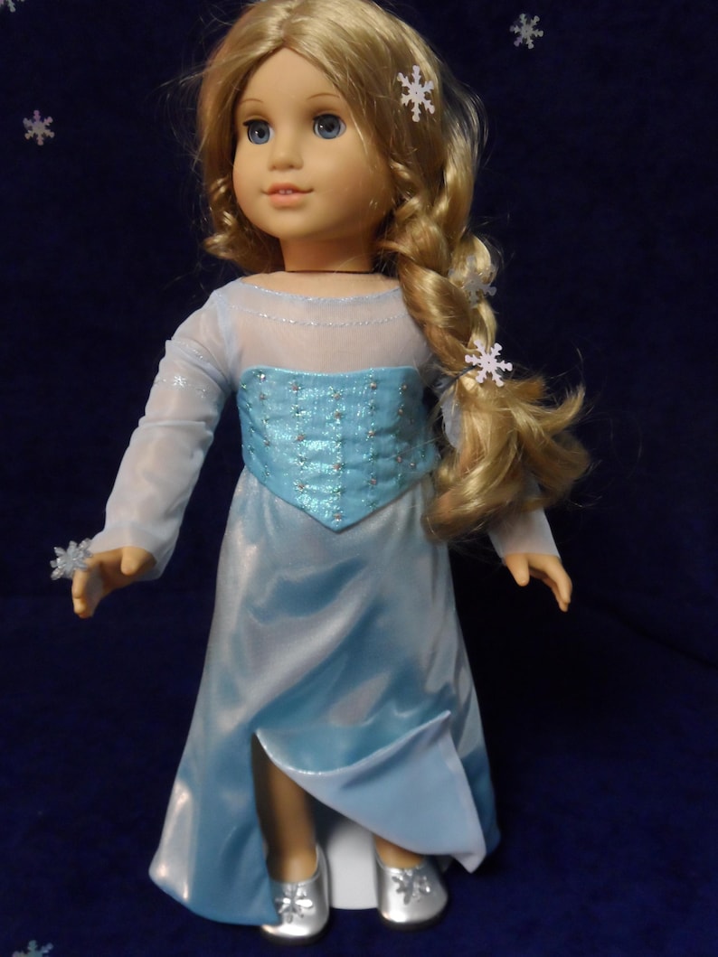 May include: A doll with blonde hair styled in a braid, wearing a light blue dress with a bodice and a slit in the skirt. The doll has blue eyes and is wearing silver shoes. Snowflake hair accessories complete the look.