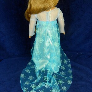 May include: A doll wearing a light blue dress with a sheer overlay and snowflake patterns. The dress has long sleeves and a flowing skirt. The doll has long blonde hair and is posed against a dark blue background.