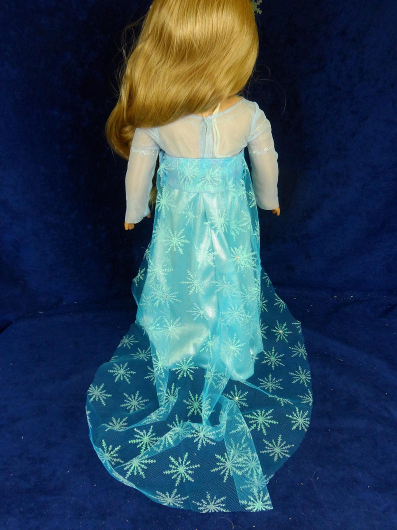 May include: A doll wearing a light blue dress with a sheer overlay and snowflake patterns. The dress has long sleeves and a flowing skirt. The doll has long blonde hair and is posed against a dark blue background.