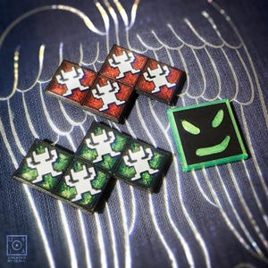 Può includere: Una collezione di tessere di gioco quadrate e colorate. Alcune tessere sono rosse con una figura bianca, altre verdi con una figura bianca, e una è nera con una faccia verde. Le tessere sono disposte su una superficie blu a motivi.