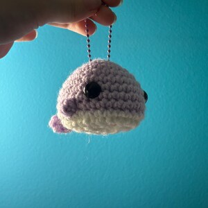 May include: A crocheted whale keychain in shades of lavender and white, featuring black button eyes. The handmade whale is suspended by a silver chain against a blue background. The whale has a small tail and is held by a hand.