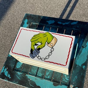 May include: A stack of white notepads with a red border, featuring a cartoon illustration of a green hand holding a small blue container attached to a chain. The hand has a gold cuff and black fingernails.
