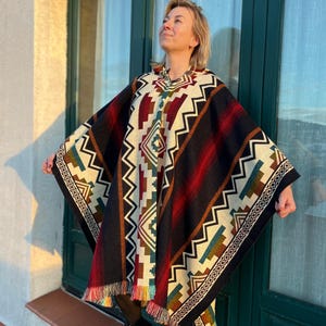 Peut inclure: Un poncho coloré avec un motif géométrique noir, rouge, crème et turquoise. Le poncho a une bordure à franges et est porté sur une tenue noire. Le design présente des zigzags et des losanges.