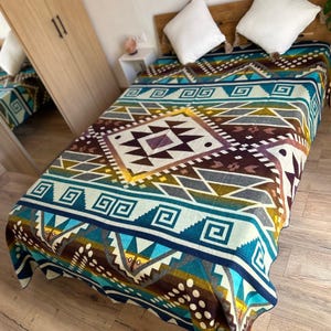 May include: A patterned blanket in teal, brown, yellow, and white covers a bed. The blanket features geometric designs, including diamond and square shapes. Two white pillows rest on the wooden headboard. The blanket is a key feature.