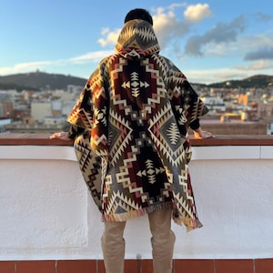 Puede incluir: Un poncho con capucha con un patrón geométrico en tonos marrones, beige, rojos y negros. El poncho tiene un dobladillo con flecos. La persona está de pie en una azotea con una vista de la ciudad al fondo.