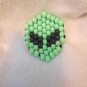 May include: A green and black beaded charm with a smiley face design. The charm is made of small, round beads that are strung together on a black thread.