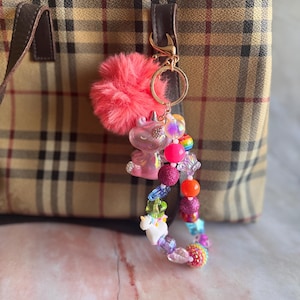 May include: A pink fluffy pom-pom keyring with a unicorn charm, iridescent beads, and a beaded bracelet with unicorn and rainbow beads. The keyring is attached to a plaid handbag with a brown leather handle.