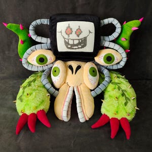 May include: A detailed plush toy depicting a fantastical creature. It features a black square head with a pixelated face, large green eyes, and multiple soft, textured appendages in various colours, including green, red, and yellow.