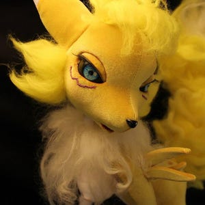 Peut inclure: Une peluche jaune avec un visage de renard, des yeux bleus et une poitrine blanche. Le jouet a une queue touffue et est fait de tissu doux. La fourrure de la créature est jaune, avec des accents blancs.