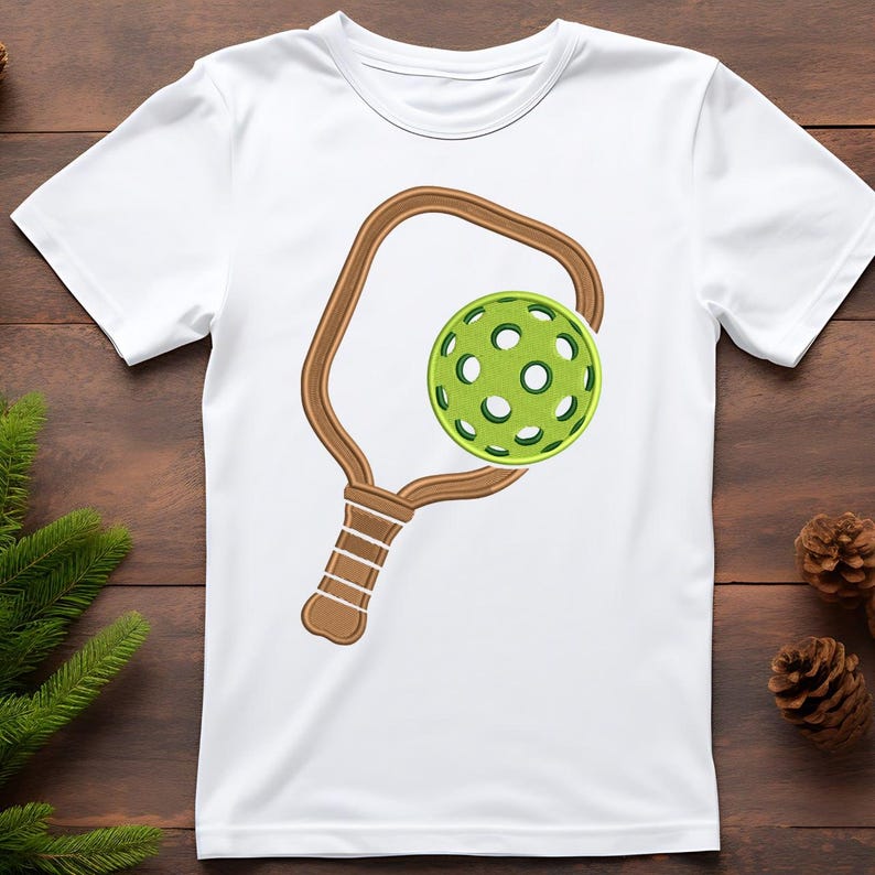 May include: White t-shirt with a brown pickleball paddle and a green pickleball design. The paddle and ball are detailed, with the ball showing a perforated pattern. The design is centered on the shirt, suitable for pickleball fans.
