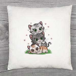 May include: White square pillow featuring a stack of three cartoon cats. The top cat is gray and white, the middle cat is gray, and the bottom cat is orange and white. Pink hearts are scattered around the cats.