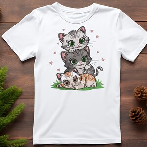 May include: White t-shirt featuring a stacked illustration of three cartoon kittens. The top kitten is gray and white, the middle one is gray, and the bottom one is orange and white. Small pink hearts are scattered around the kittens.