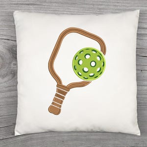 May include: White square cushion with an embroidered pickleball paddle and ball design. The paddle is brown with a textured handle, and the ball is green with circular cutouts. The design is centered on the cushion.