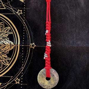 May include: A circular, pale green and gray stone pendant with a central hole, suspended by a red braided cord. The cord features silver-colored accents and a knotted top. The pendant is displayed against a dark background.