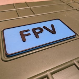 Peut inclure: Une boîte verte avec une étiquette rectangulaire bleue qui dit "FPV" en lettres noires.