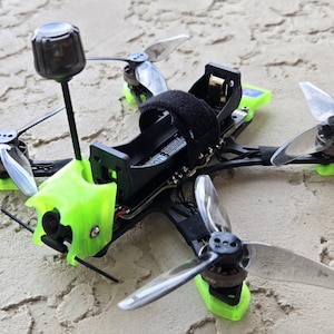 May include: A small, black and green drone with four propellers. The drone has a black body with green accents and silver propellers. A black antenna extends from the top. The drone is resting on a textured, light-colored surface.
