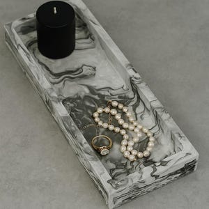 May include: A rectangular marble-effect tray with a black candle, a pearl necklace, and a gold ring. The tray has a swirling gray and white pattern. The ring has a large, clear stone.