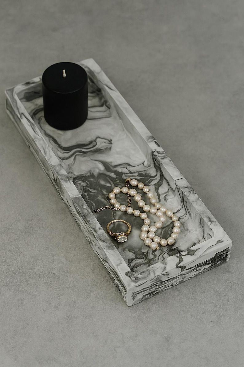 May include: A rectangular marble-effect tray with a black candle, a pearl necklace, and a gold ring. The tray has a swirling gray and white pattern. The ring has a large, clear stone.