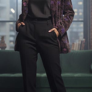 Op de afbeelding: Een vrouw draagt een zwarte top en broek, met een bloemenblazer in paarse en bruine tinten. Ze draagt zwarte hakken. De achtergrond bevat een groene bank en een raam.