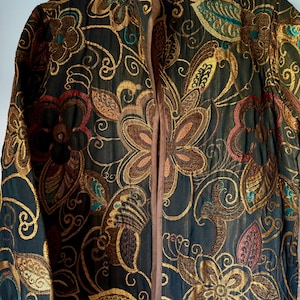 May include: A long-sleeved jacket featuring a dark base with intricate, multi-colored floral embroidery in shades of gold, copper, red, and teal. The garment has a high collar, a central vertical opening, and brown trim along the edges.