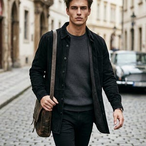 May include: A person wearing a black suede jacket over a grey jumper and black trousers, carrying a brown leather messenger bag. The person is walking on a cobbled street.