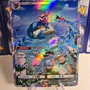 May include: A holographic Vaporeon Pok&eacute;mon trading card. The card features a blue and pink Vaporeon character in a water-themed scene. The card has text that reads "Vaporeon", "Wash Out", and "Wave Splash".