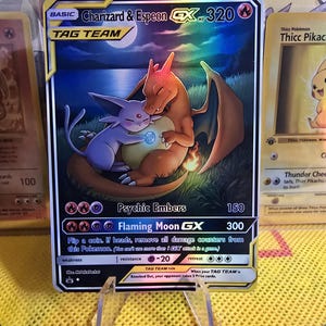 May include: A Charizard & Espeon GX Tag Team trading card. The card features a Charizard and Espeon embracing under a full moon. The card has a HP of 320 and includes the text "Psychic Embers" and "Flaming Moon GX".