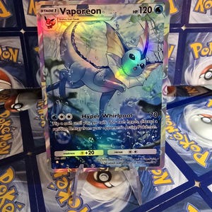 May include: A holographic Vaporeon Pok&eacute;mon card. The card features a blue and yellow Vaporeon character with the text "Vaporeon" and "HP 120". The card is displayed on a clear plastic stand.