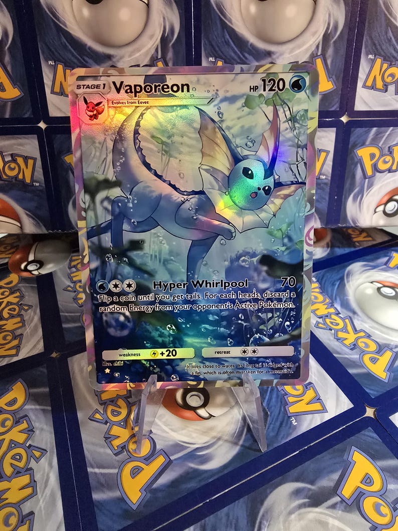 May include: A holographic Vaporeon Pok&eacute;mon card. The card features a blue and yellow Vaporeon character with the text "Vaporeon" and "HP 120". The card is displayed on a clear plastic stand.