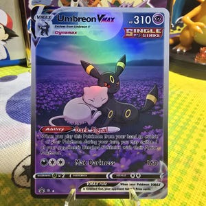 Puede incluir: Una carta Pokémon Umbreon VMAX con Umbreon y Mew abrazándose en un campo de flores moradas. La carta tiene un diseño Single Strike y dice "Max Darkness 160". La carta se muestra en un soporte transparente.