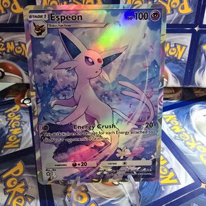 May include: A holographic Pok&eacute;mon card featuring Espeon, a Stage 1 Pok&eacute;mon. The card has a HP of 100 and includes the text "Energy Crush". The background shows a cloudy sky with pink and purple accents. The card is displayed on a clear stand.