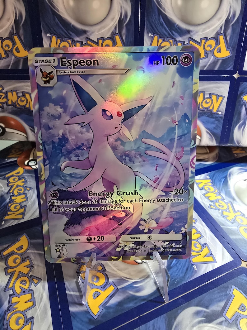 May include: A holographic Pok&eacute;mon card featuring Espeon, a Stage 1 Pok&eacute;mon. The card has a HP of 100 and includes the text "Energy Crush". The background shows a cloudy sky with pink and purple accents. The card is displayed on a clear stand.