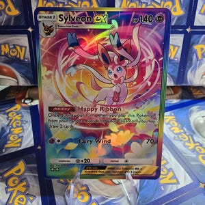 May include: A holographic Pok&eacute;mon card featuring Sylveon EX. The card has a colorful background with a pink and white Sylveon character. The card text includes "Happy Ribbon" and "Fairy Wind". The card is displayed on a clear stand.