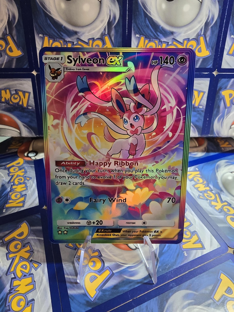 May include: A holographic Pok&eacute;mon card featuring Sylveon EX. The card has a colorful background with a pink and white Sylveon character. The card text includes "Happy Ribbon" and "Fairy Wind". The card is displayed on a clear stand.