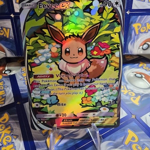 May include: A holographic Eevee EX Pok&eacute;mon trading card. The card features a colorful illustration of Eevee surrounded by flowers and foliage. The card has text that includes "Basic", "HP 90", "Veevee Volve", and "Bite".