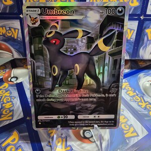 May include: A holographic Umbreon Pok&eacute;mon card. The card features a black Umbreon with yellow accents and red eyes. The card has the text "Umbreon" and "Dark Binding" printed on it. The card is displayed on a clear stand.