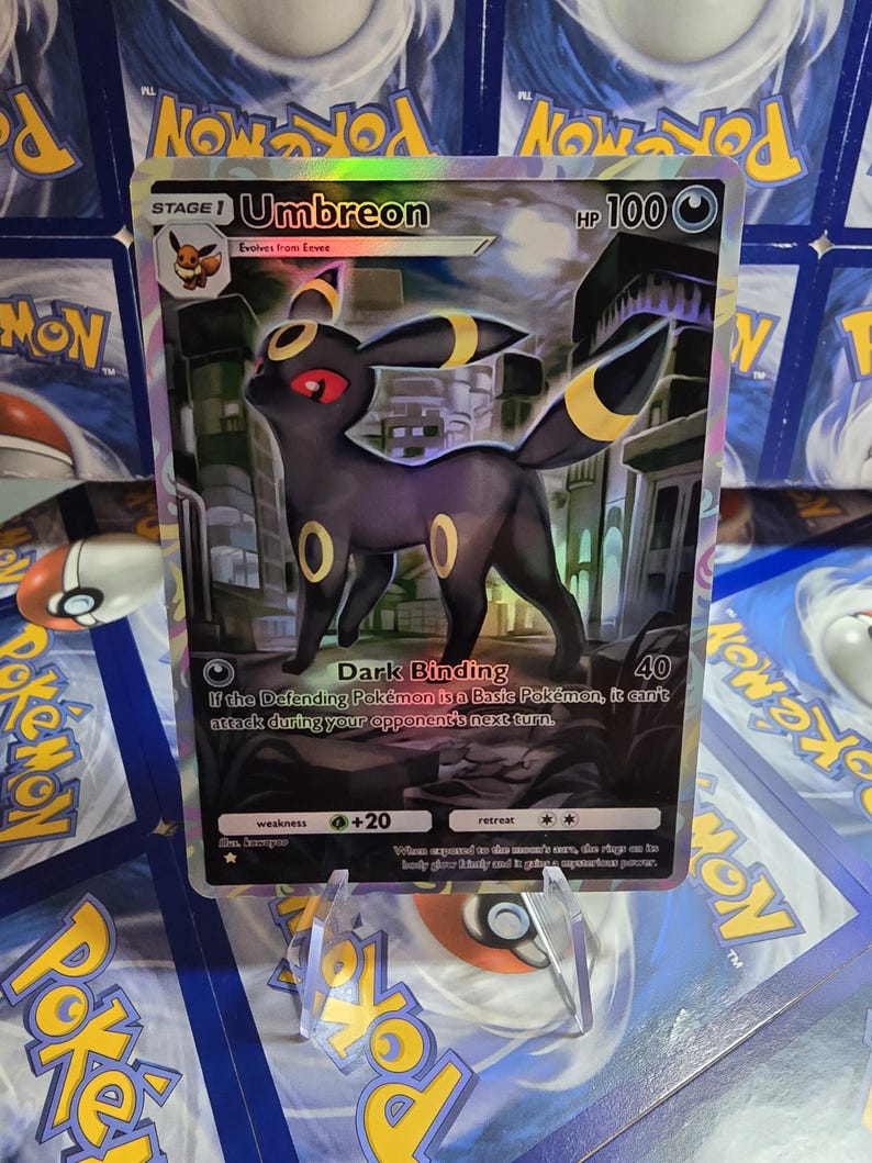 May include: A holographic Umbreon Pok&eacute;mon card. The card features a black Umbreon with yellow accents and red eyes. The card has the text "Umbreon" and "Dark Binding" printed on it. The card is displayed on a clear stand.