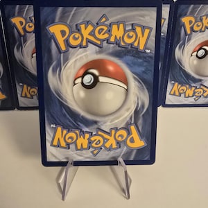 May include: A blue-backed Pok&eacute;mon trading card featuring the Pok&eacute;mon logo and a Pok&eacute; Ball design. The card is displayed on a clear plastic stand, with other cards visible in the background.