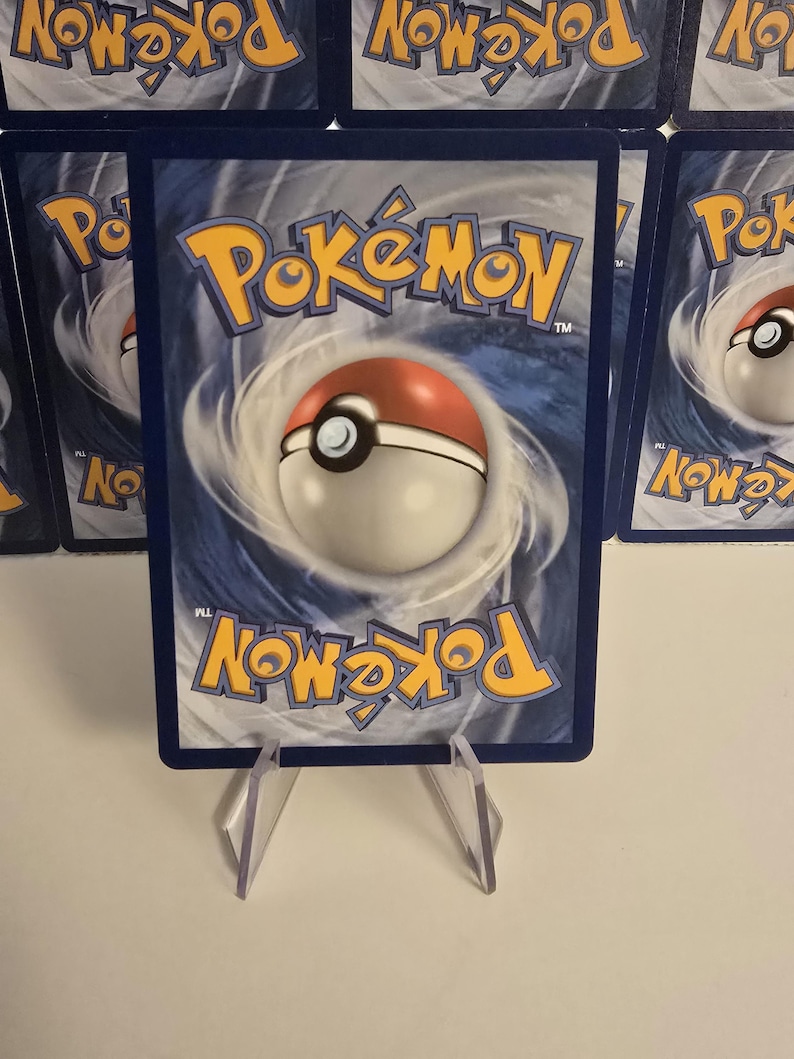 May include: A blue-backed Pok&eacute;mon trading card featuring the Pok&eacute;mon logo and a Pok&eacute; Ball design. The card is displayed on a clear plastic stand, with other cards visible in the background.