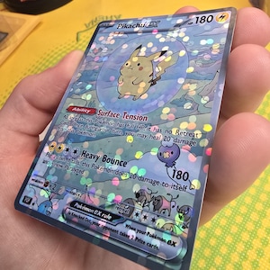K&ouml;nnte beinhalten: Holografische Pikachu EX Pok&eacute;mon-Sammelkarte. Die Karte zeigt einen gelben Pikachu-Charakter in einer Blase, mit Text, der seine F&auml;higkeiten und Werte beschreibt. Die Karte hat einen silbernen Rand und wird in einer Hand gehalten.