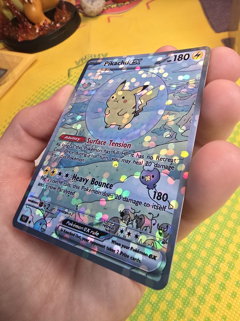K&ouml;nnte beinhalten: Holografische Pikachu EX Pok&eacute;mon-Sammelkarte. Die Karte zeigt einen gelben Pikachu-Charakter in einer Blase, mit Text, der seine F&auml;higkeiten und Werte beschreibt. Die Karte hat einen silbernen Rand und wird in einer Hand gehalten.