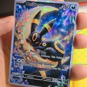 K&ouml;nnte beinhalten: Holografische Umbreon Pok&eacute;mon-Sammelkarte. Die Karte zeigt einen schwarz-gelben Umbreon-Charakter vor einem Vollmond. Die Karte hat einen blau-violetten Sternenhintergrund mit den Texten "Moonlit Nap" und "Shattering Slumber".