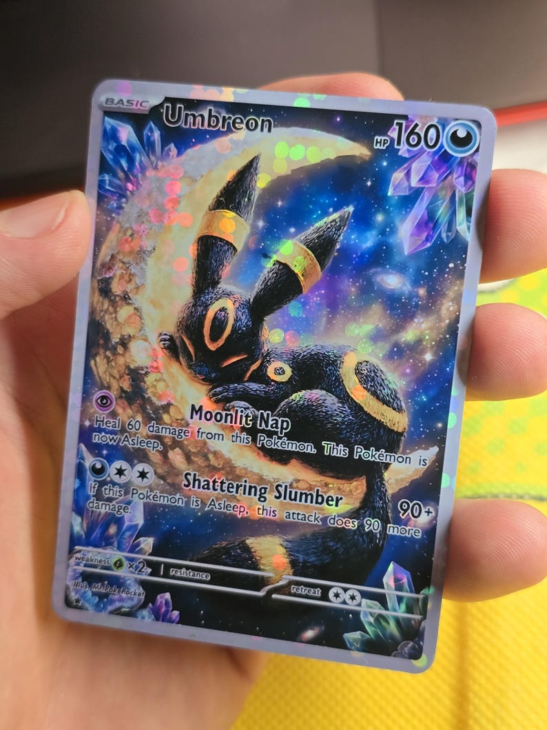 K&ouml;nnte beinhalten: Holografische Umbreon Pok&eacute;mon-Sammelkarte. Die Karte zeigt einen schwarz-gelben Umbreon-Charakter vor einem Vollmond. Die Karte hat einen blau-violetten Sternenhintergrund mit den Texten "Moonlit Nap" und "Shattering Slumber".