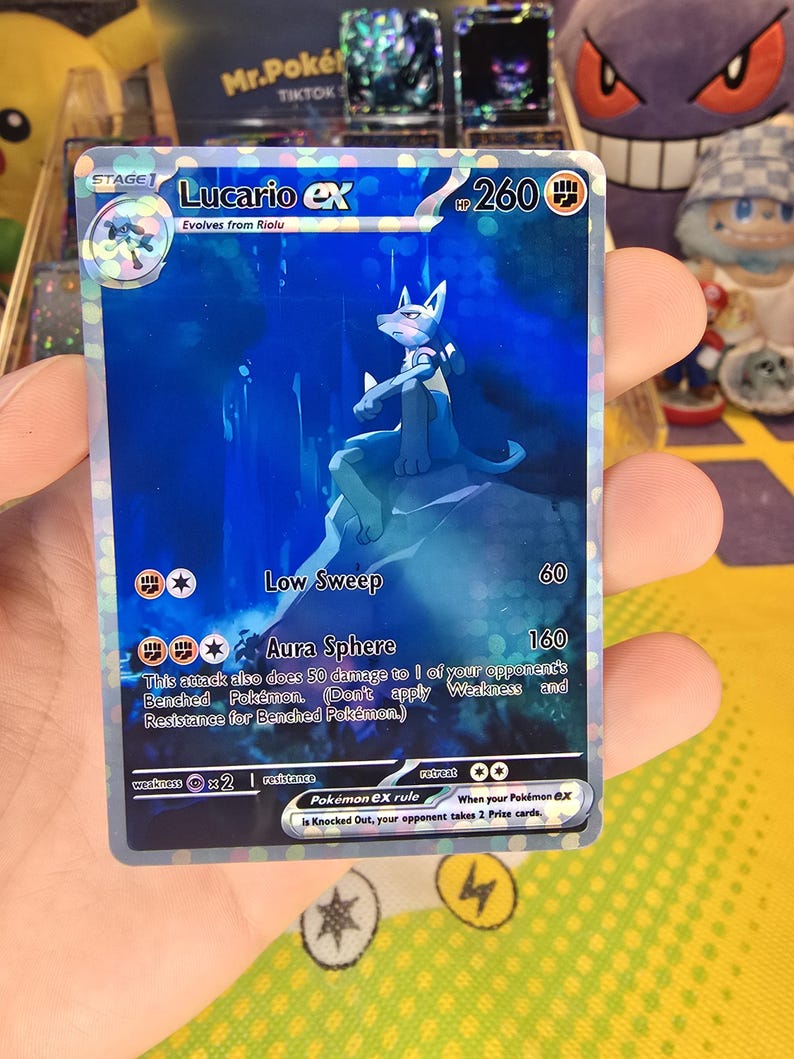 K&ouml;nnte beinhalten: Eine holografische Lucario EX Pok&eacute;mon-Sammelkarte. Die Karte zeigt einen blauen Lucario-Charakter mit dem Text "Lucario EX" und "Aura Sphere". Die Karte hat 260 HP und den Text "Evolves from Riolu".