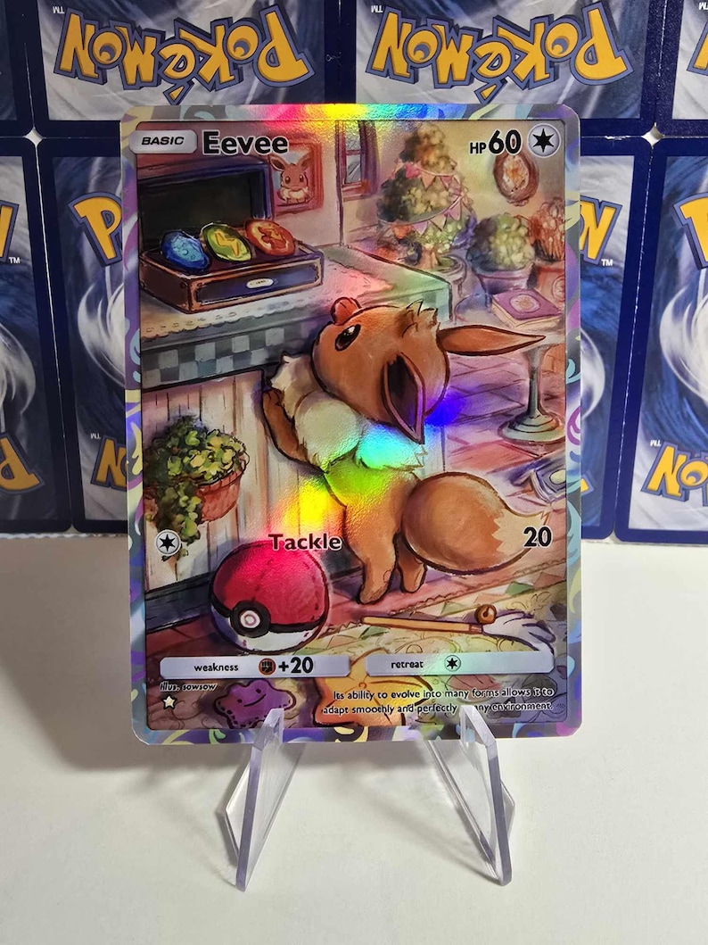 May include: A holographic Eevee Pok&eacute;mon trading card. The card features an illustration of Eevee in a room with a Pok&eacute; Ball and text that reads "Tackle" and "HP 60". The card is displayed on a clear stand.