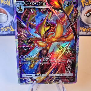 May include: A holographic Alakazam Pok&eacute;mon trading card. The card features a vibrant illustration of Alakazam in shades of yellow, orange, and blue. The card text includes "Alakazam", "Psychic", and "HP 130".