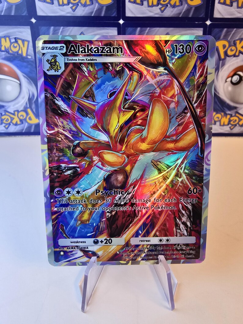 May include: A holographic Alakazam Pok&eacute;mon trading card. The card features a vibrant illustration of Alakazam in shades of yellow, orange, and blue. The card text includes "Alakazam", "Psychic", and "HP 130".