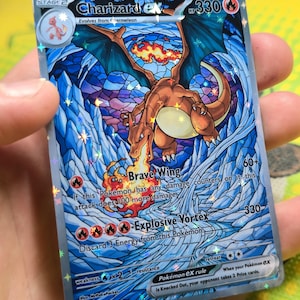 K&ouml;nnte beinhalten: Eine Charizard EX Pok&eacute;mon-Sammelkarte. Die Karte zeigt eine lebendige Illustration von Charizard mit blauen Fl&uuml;geln und einem feurigen Schwanz, vor einem Hintergrund im Buntglasstil. Die Karte hat den Text "Brave Wing" und "Explosive Vortex".