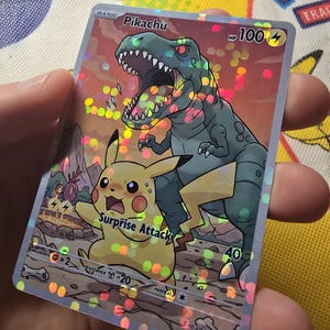 May include: A holographic Pikachu trading card featuring a yellow Pikachu character and a green dinosaur. The card has the text "Surprise Attack" and "HP 100". The card is held in a person's hand.